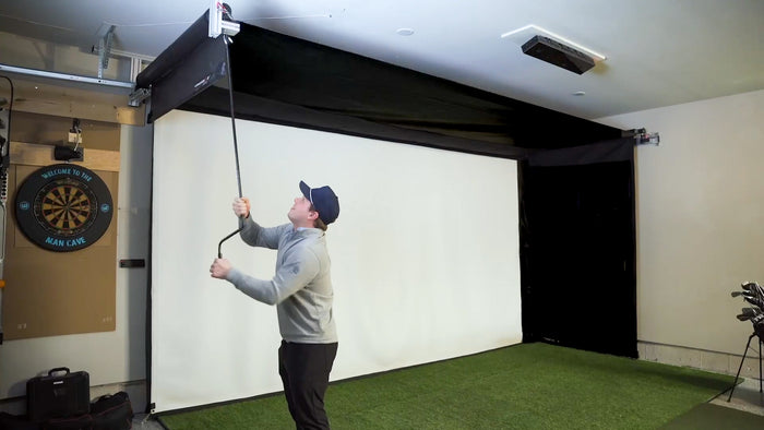 Man operating a golf simulator side screen using hand crank in a room with a screen and golf equipment.