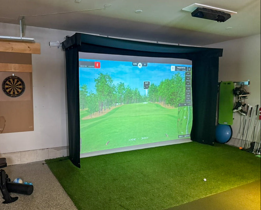 Indoor golf simulator setup with screen displaying a golf course, chair, and various equipment.