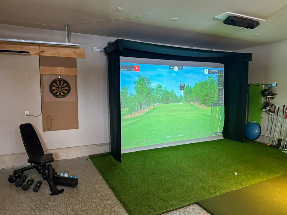 Indoor golf simulator setup with screen displaying a golf course, chair, and various equipment.