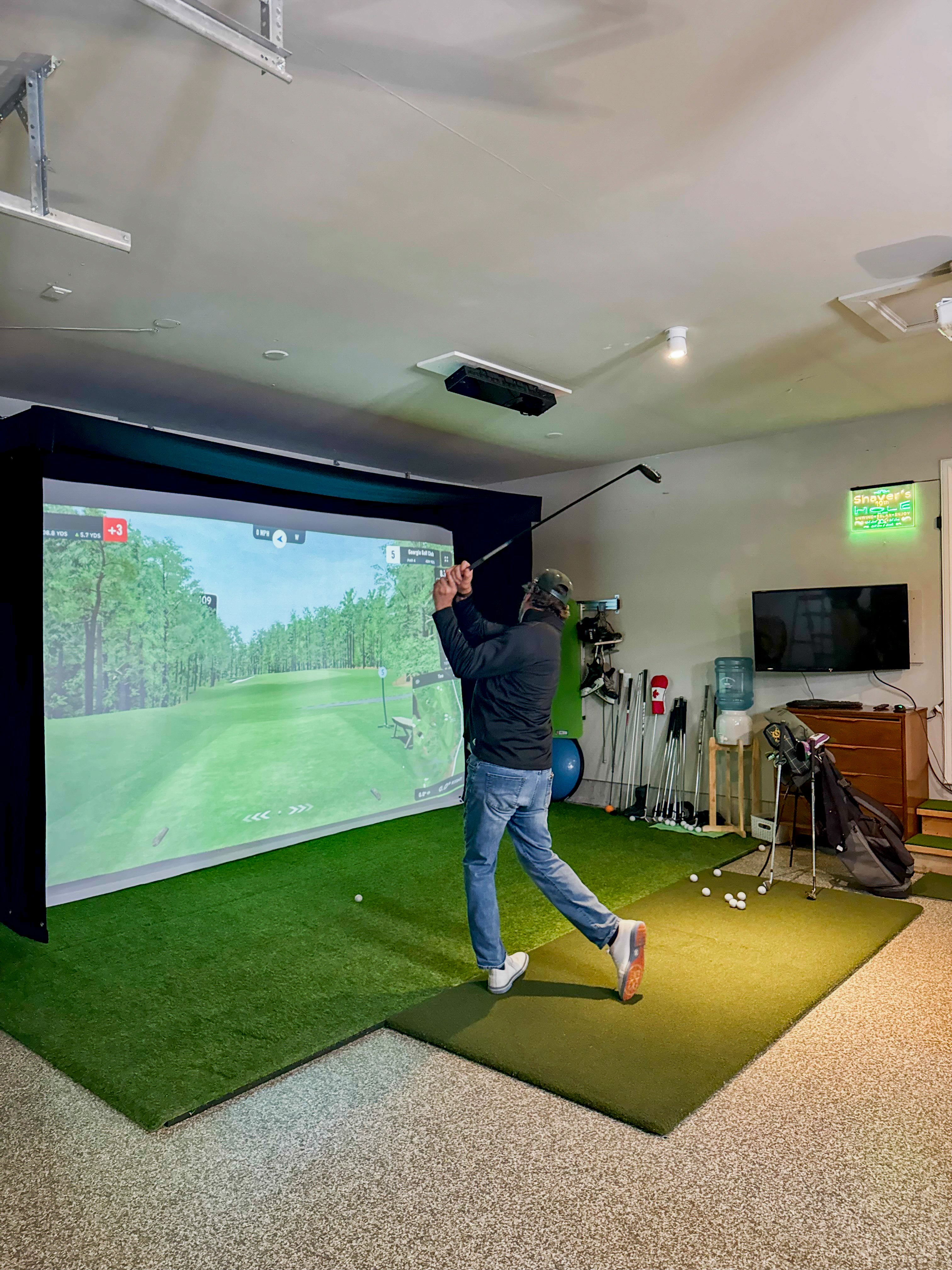 Person playing golf simulator in a room with a large screen and golf equipment.